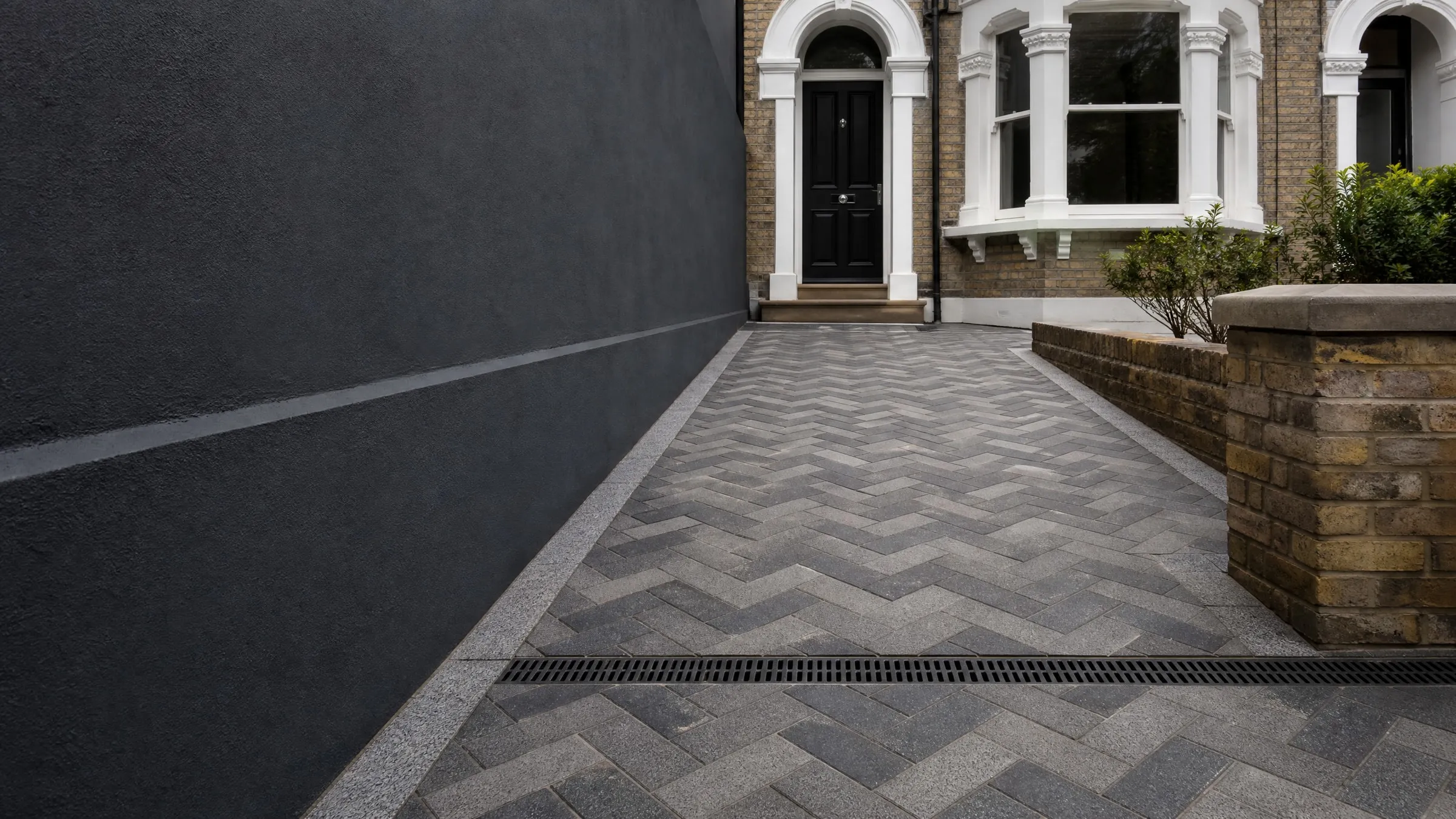 Charcoal herringbone block paving with granite edging and a drainage channel leading to a Victorian front door
